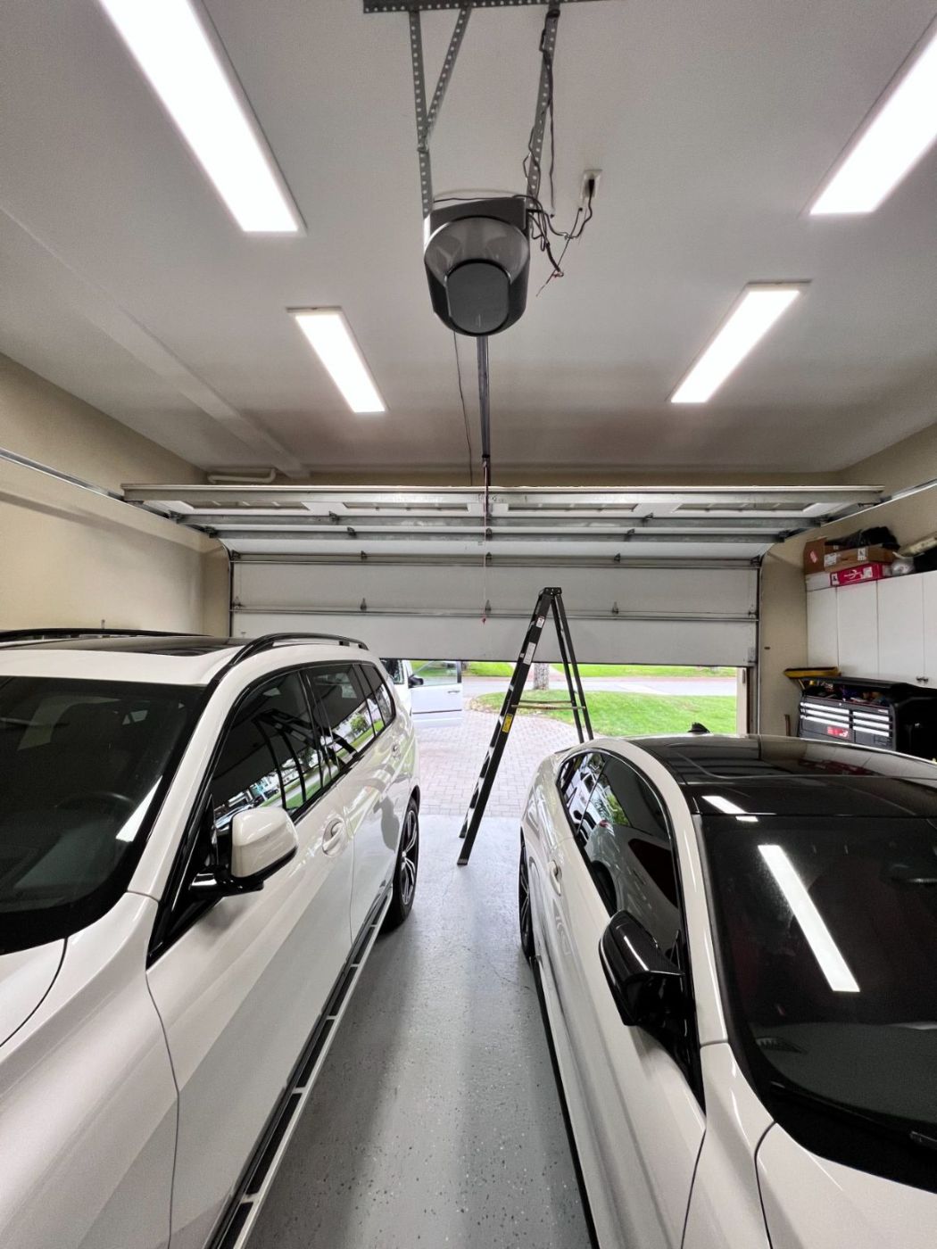 Clean modern Coquitlam garage interior with two white SUVs parked under a freshly serviced wall-mount jackshaft opener, ladder set up under the opener for a service call