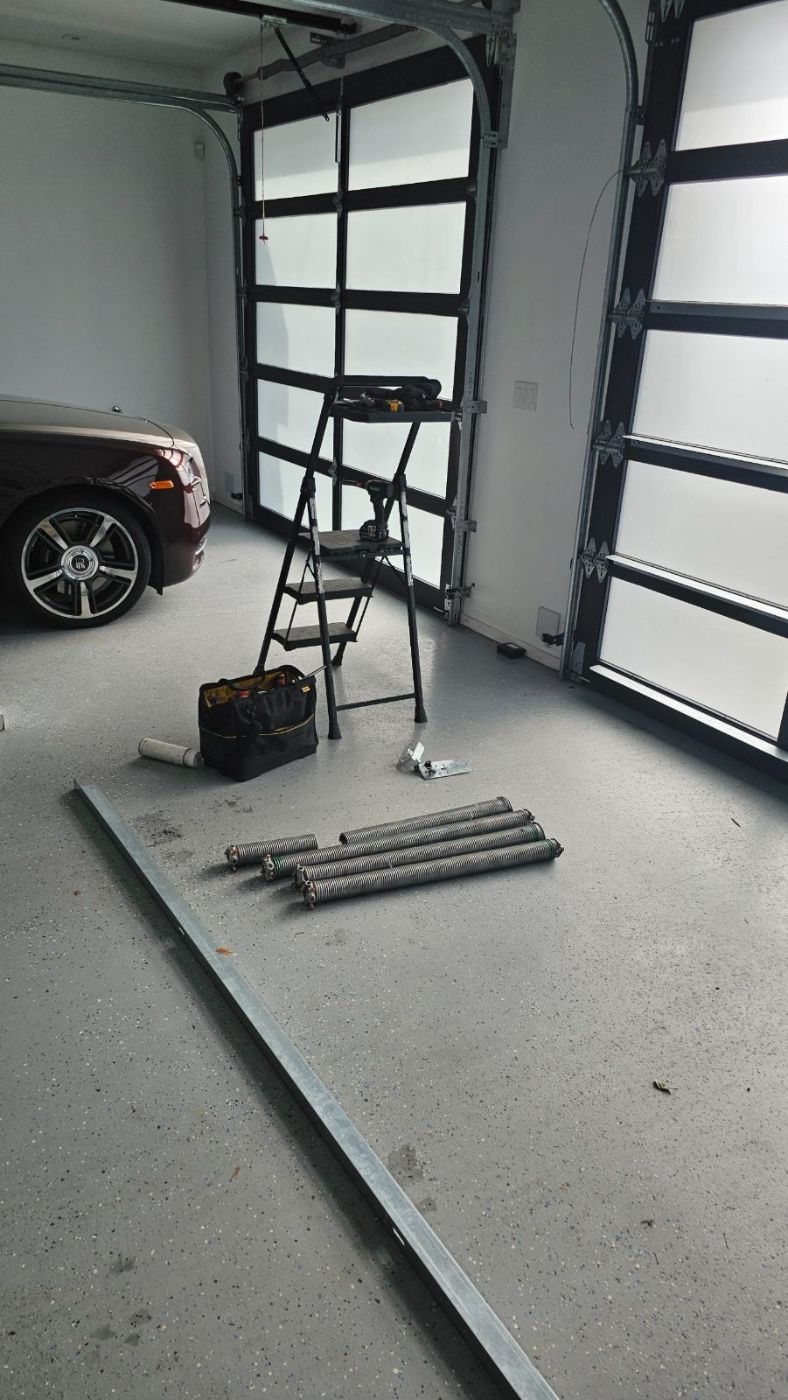 Four new heavy-duty galvanized torsion springs lined up on the floor of a high-end Coquitlam garage with a Rolls-Royce parked beside them, ready for installation