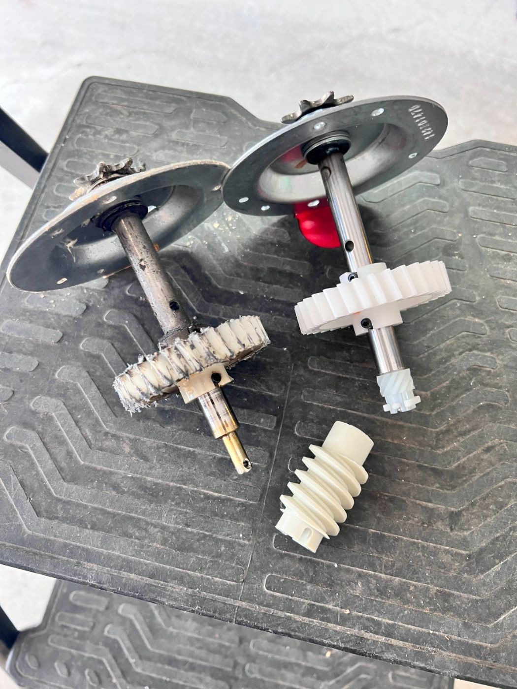 Old chewed-up LiftMaster drive gear and sprocket assembly next to a brand-new white nylon replacement gear kit on a Port Moody work bench
