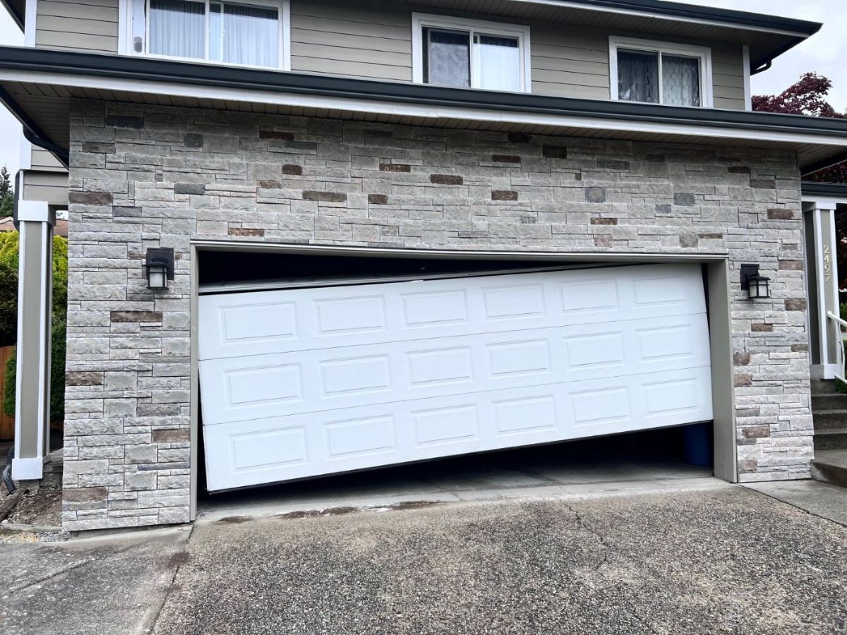 White sectional garage door knocked off track on a Port Moody home with stone facade, panels hanging at an angle in the opening