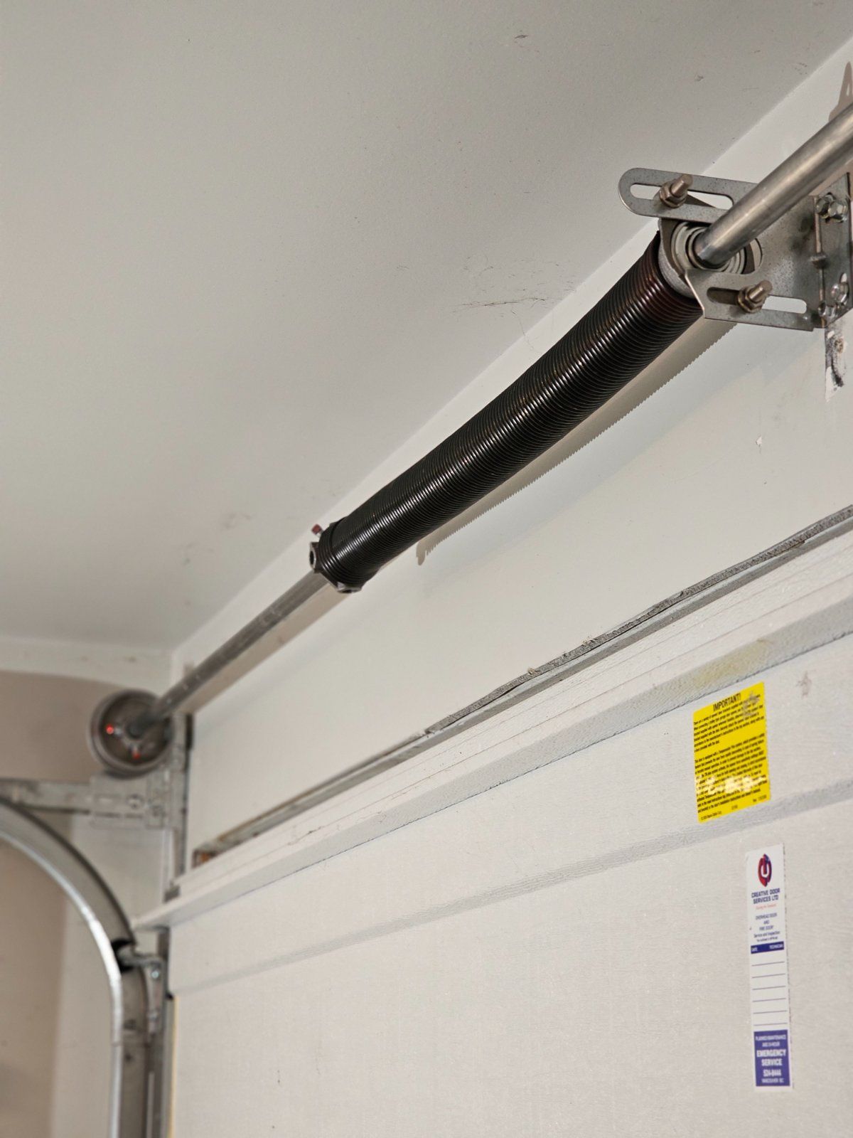 Black torsion spring mounted on a clean shaft above a residential garage door in Port Moody after a preventive maintenance service visit, with Creative Door service tag visible