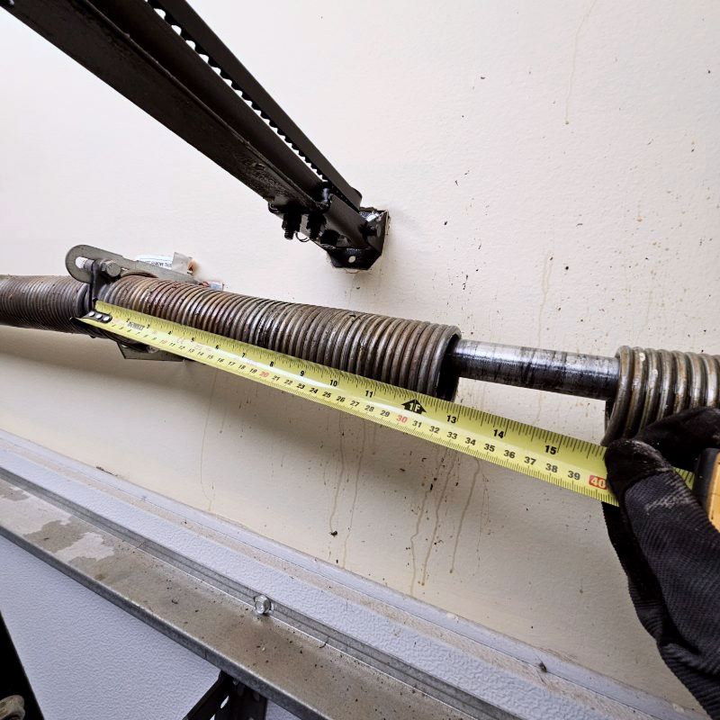 Technician measuring a broken garage door torsion spring with a tape measure to determine correct replacement size